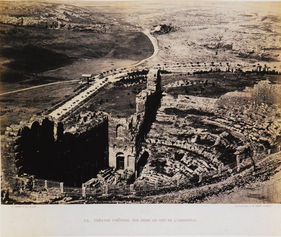 The Odeion of Herodes Atticus amidst a treeless landscape and Loumbardiaris to the far back amidst green; the two main roads join as they do nowadays (The Gennadius Library - American School of Classical Studies at Athens, from the website of the Aikaterini Laskaridi Foundation).